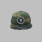 A camouflage, flat bill, snapback equality hat with a black round patch, outlined by white. The embroidered text on the patech says Fifty50, Equality, in white, with two Fs, back to back, in the middle.