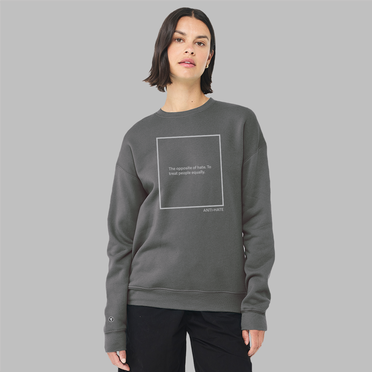 A lady wearing a charcoal grey unisex sweatshirt with a light grey logo. The text, inside a large rectangle, reads, "The opposite of hate. To treat people equally." Anti-hate is written in the lower left corner. 