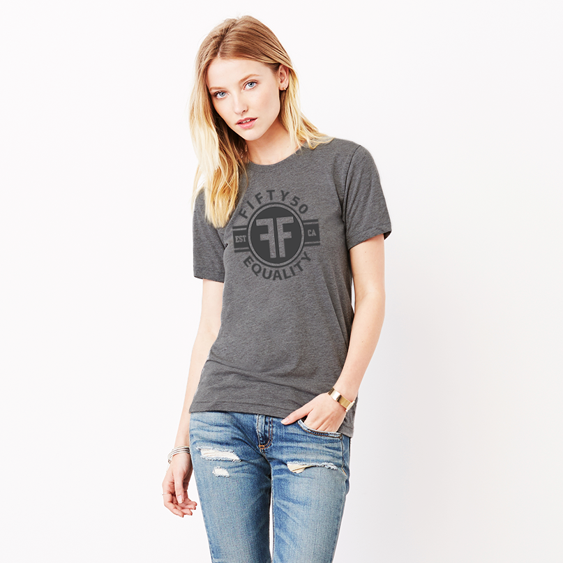 A lady wearing a charcoal grey equality tshirt with a black graphic.  The logo text says Fifty50, Equality, and there are two Fs, back to back in the center inside a circle. 