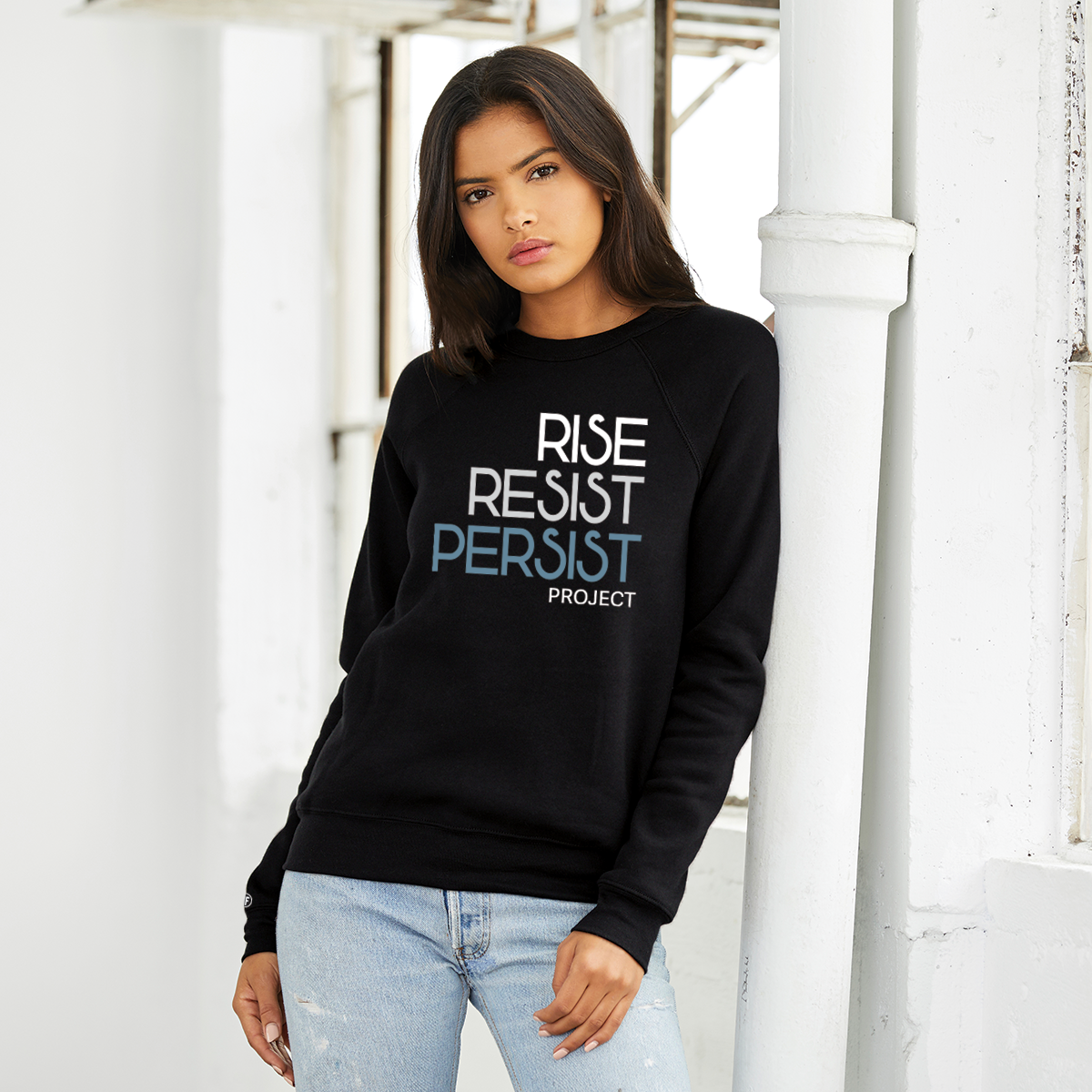 A lady wearing a political unisex black sweatshirt. The text says Rise, Resist, Persist, Project. The font is white, Slate Blue, and grey.